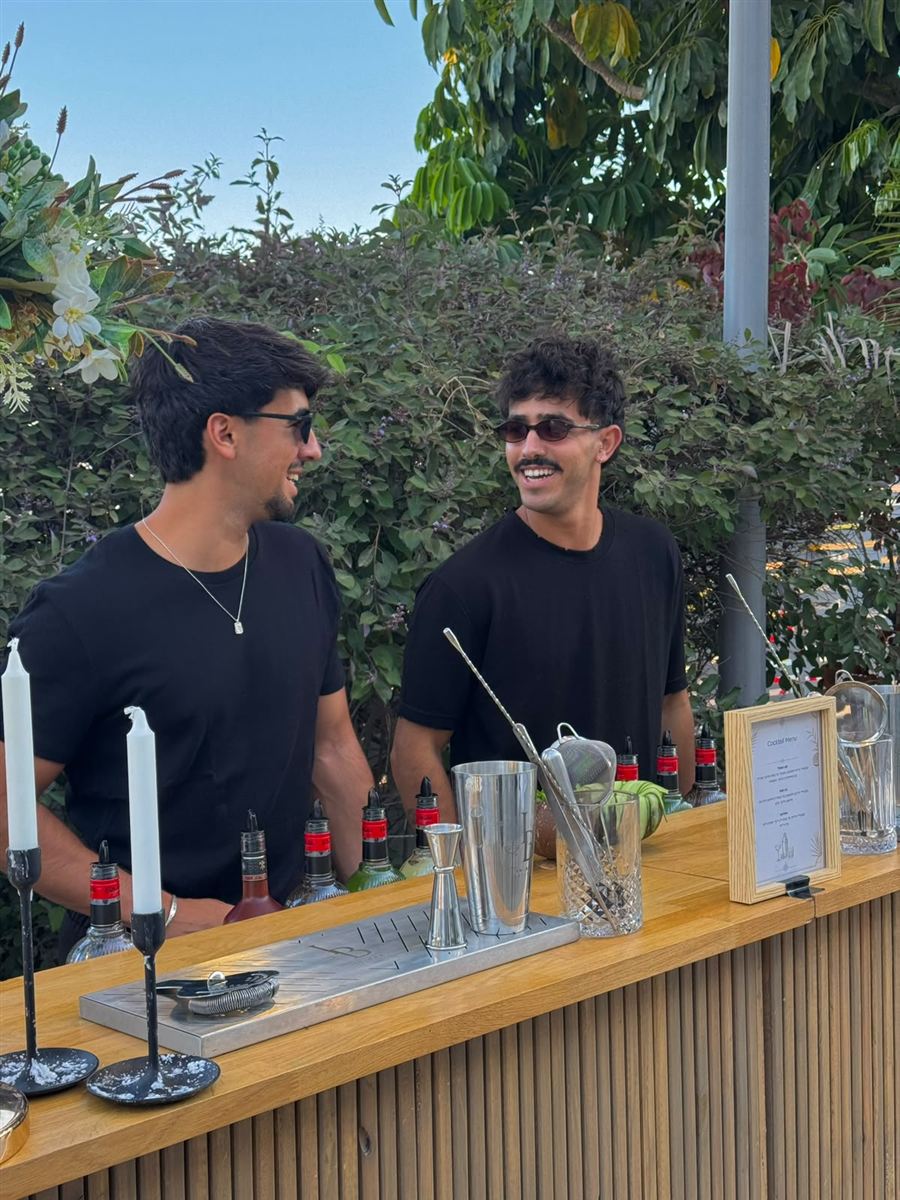 שני ברמנים של IN BAR צוחקים מאחורי בר עץ עם נרות ותפריט קוקטיילים