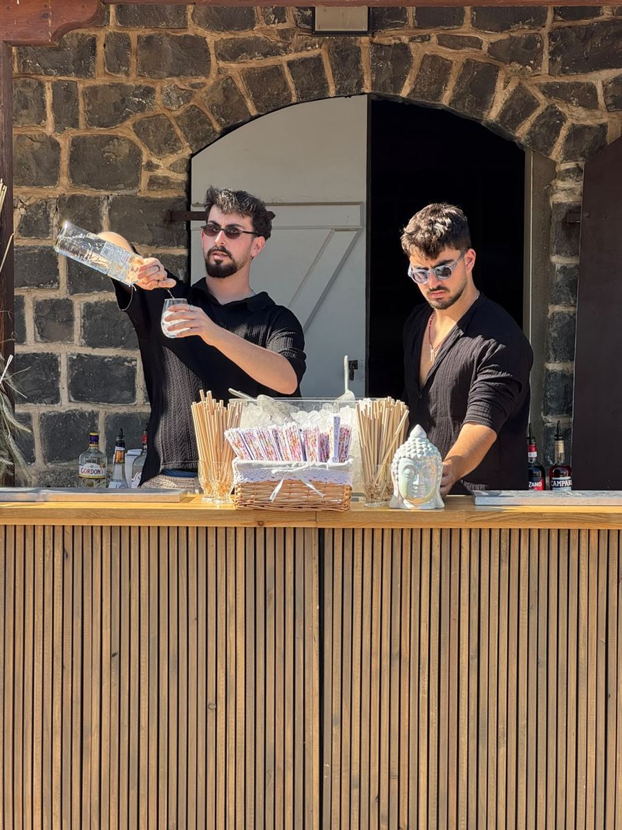 שני ברמנים של IN BAR מוזגים משקאות בבר עץ מול קיר אבן