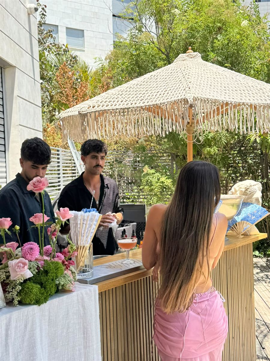 בר עץ של IN BAR מתחת לשמשיית מקרמה, עם פרחים ורודים ואורחת ליד הבר
